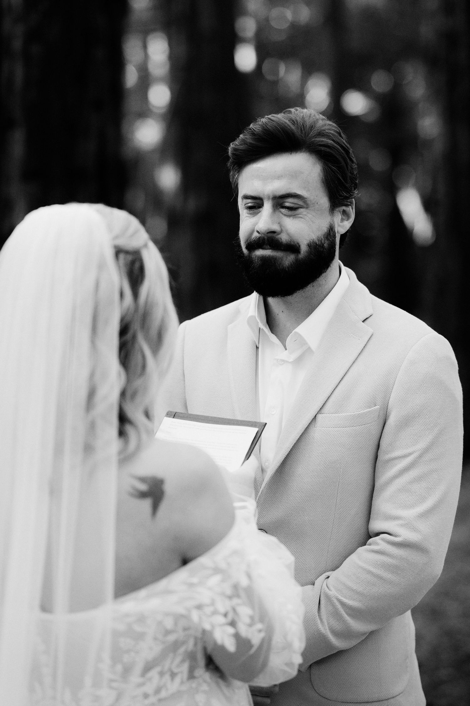 A black and white photo of a wedding ceremony outdoors, showing a bearded groom in a light suit reading vows.
