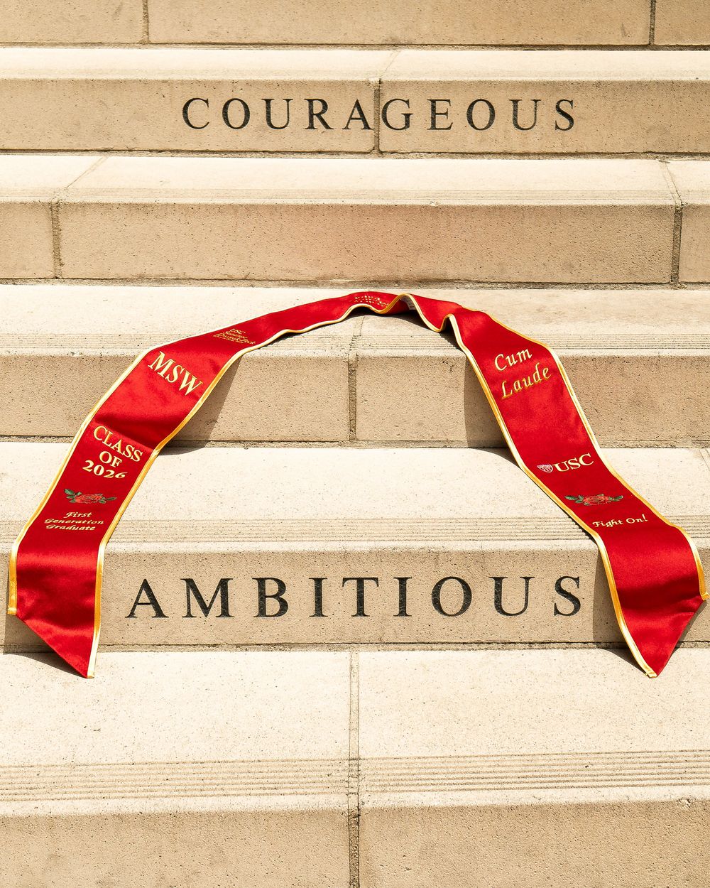 Red graduation stole draped across campus steps engraved with words “Courageous” and “Ambitious,” symbolizing college graduate success.