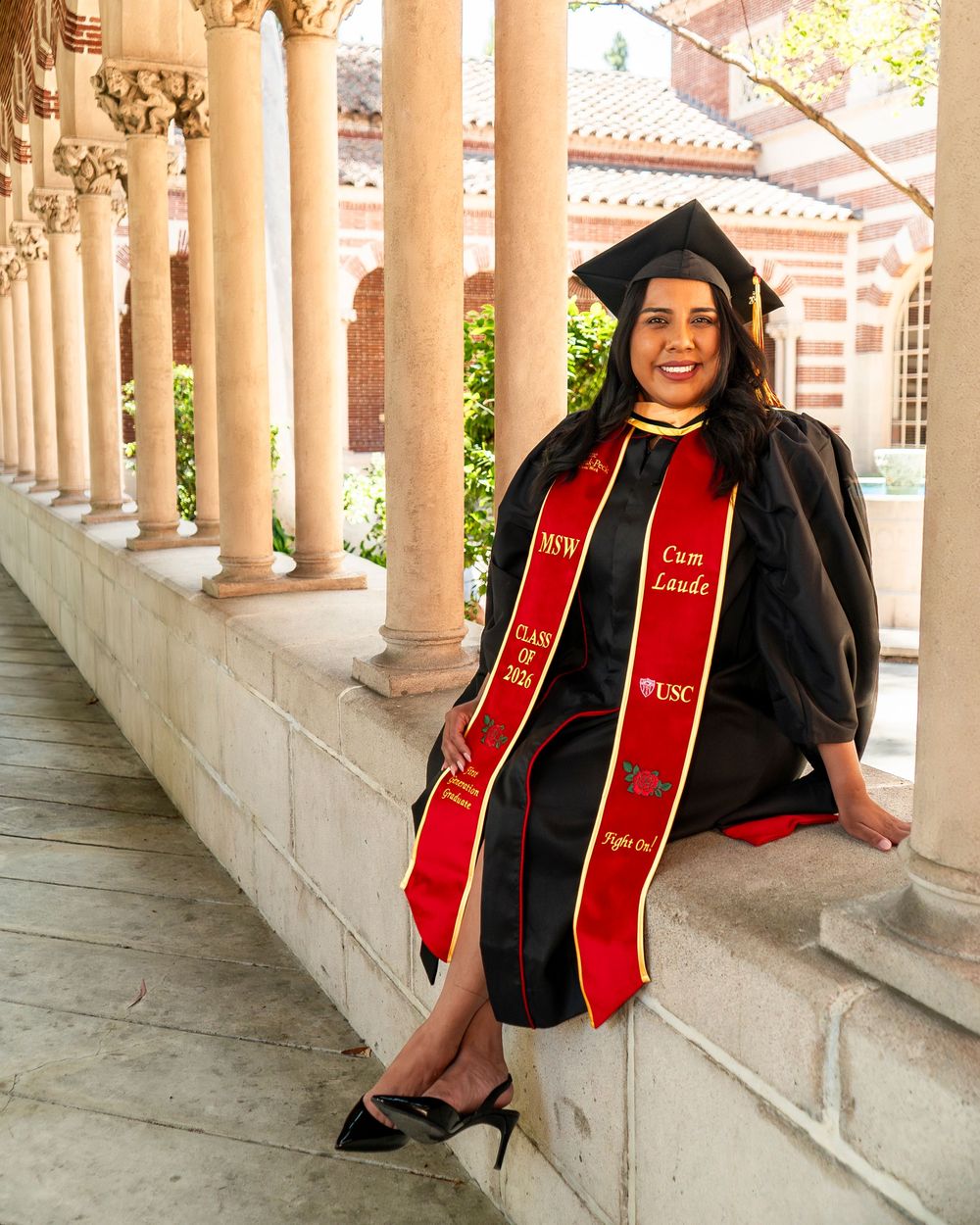 Woman in cap and gown with red honor stoles sitting along columned hallway, proud university graduate posing on campus.
