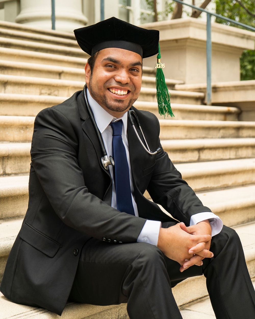 Graduate in suit with cap and stethoscope sitting on campus steps, celebrating new medical school graduation and healthcare career.