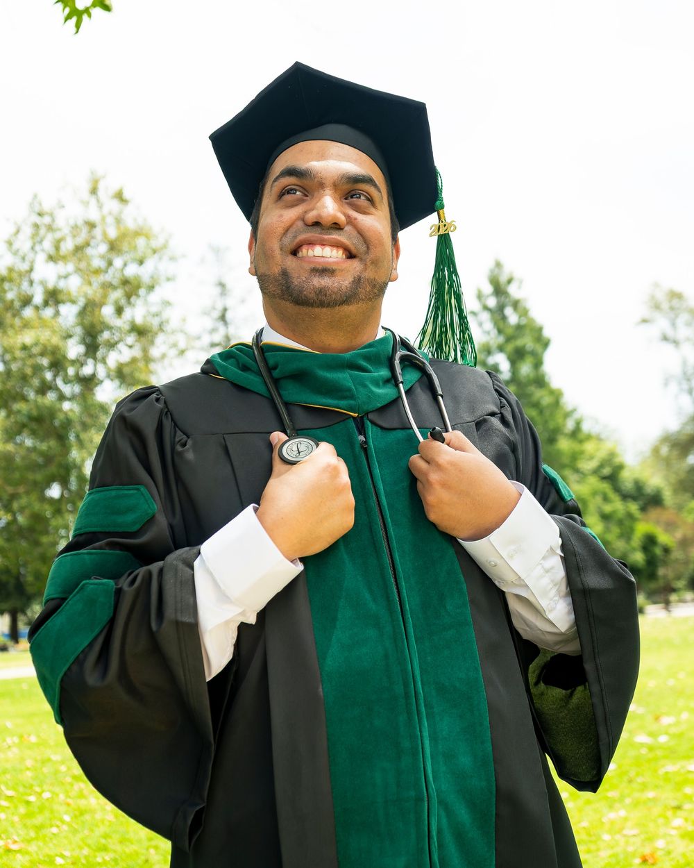 Medical graduate wearing green and black doctoral gown with stethoscope, posing proudly outdoors after graduation ceremony.
