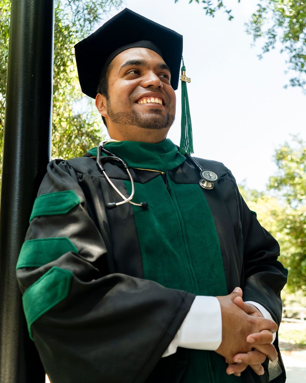 Confident medical school graduate in doctoral regalia with stethoscope, standing outside on campus on graduation day.