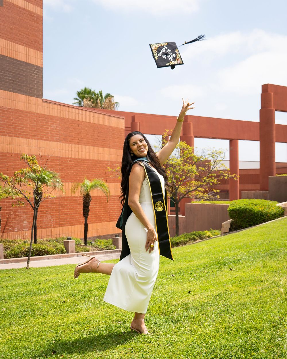 Graduate in white dress and stole tosses decorated cap in the air on a grassy campus hill.