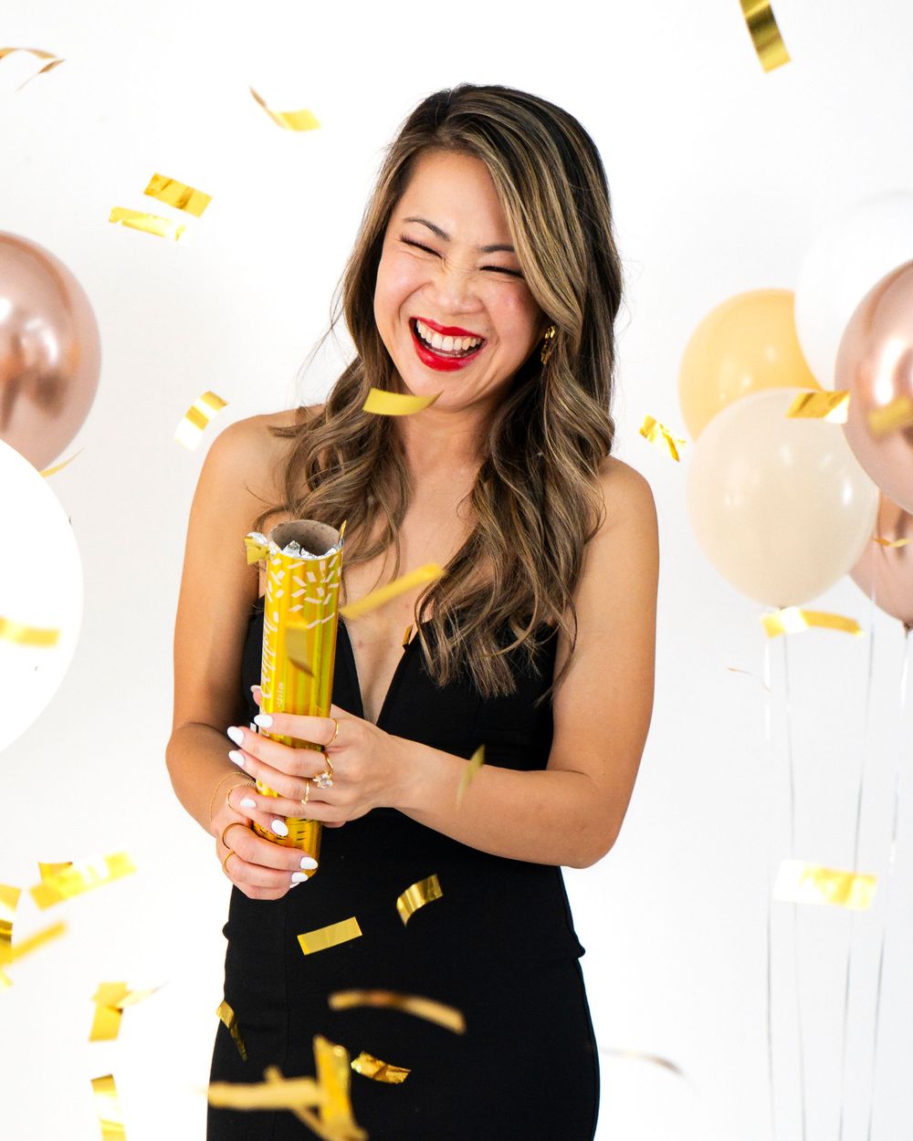 Woman in black dress popping a gold confetti cannon surrounded by balloons.
