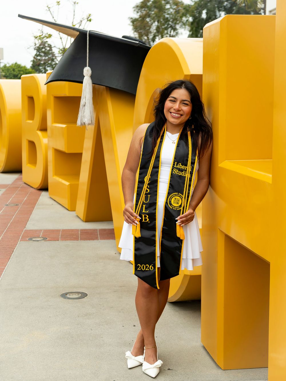 College graduate in white dress and black stole posing by large yellow campus letters, celebrating university graduation and class of 2026.