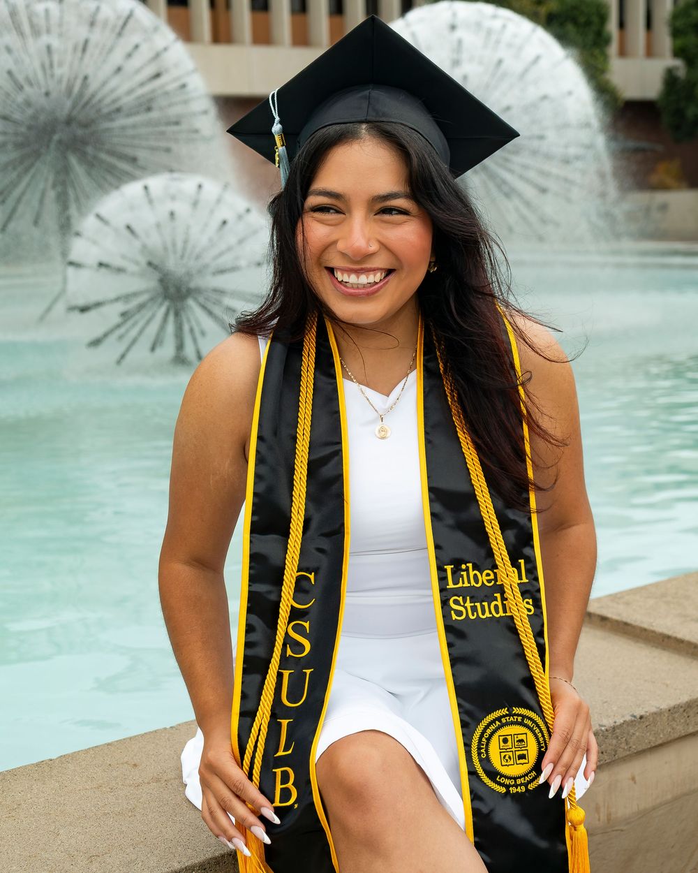 Graduate in white dress and cap sitting by a fountain, wearing a black and gold CSULB Liberal Studies stole.