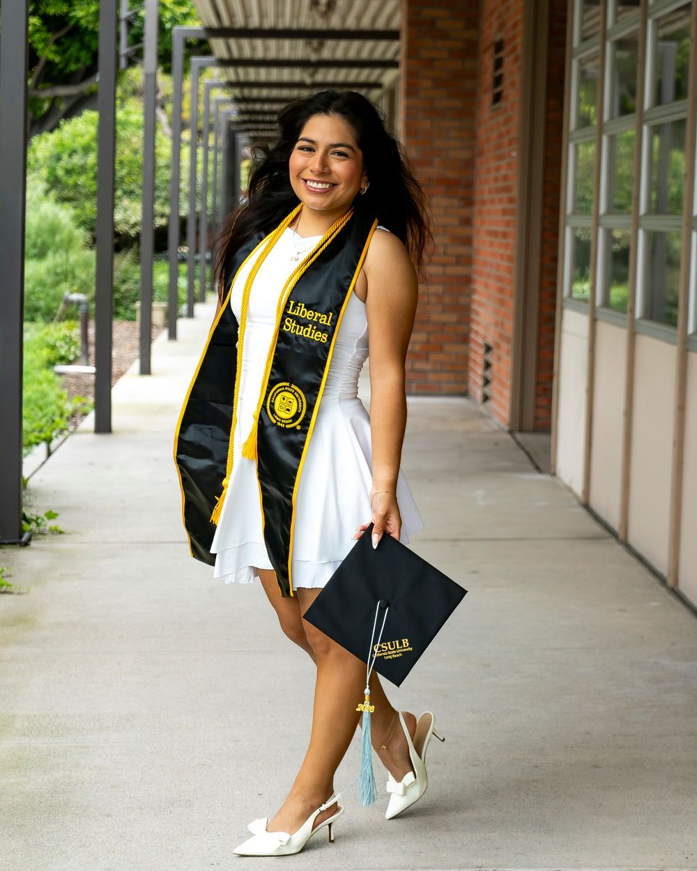 Graduate in white dress and heels under a campus walkway, holding a graduation cap and wearing a black and gold stole.