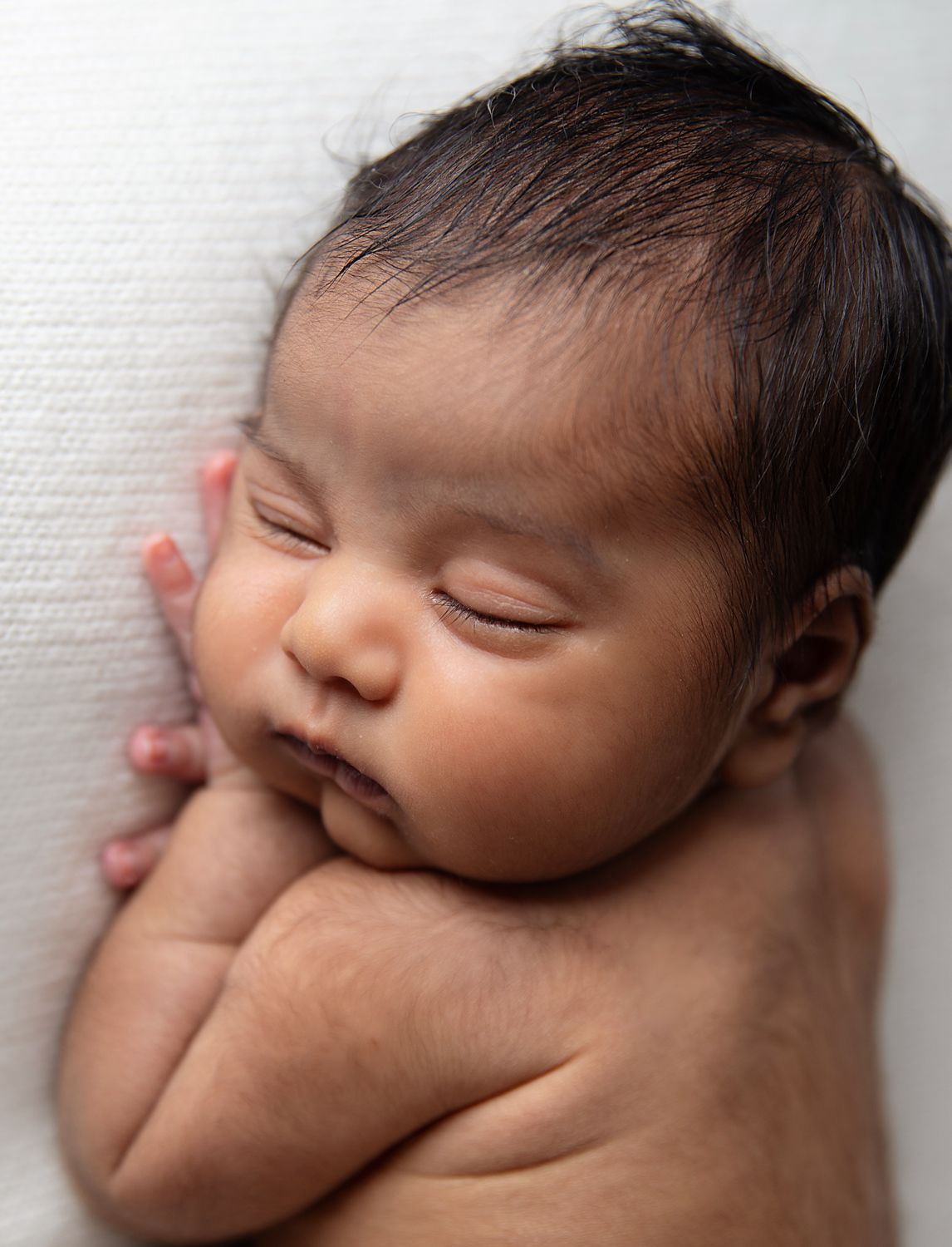 Peaceful sleeping newborn baby with dark hair lies curled up against white textured background. During a newborn photoshoot in London