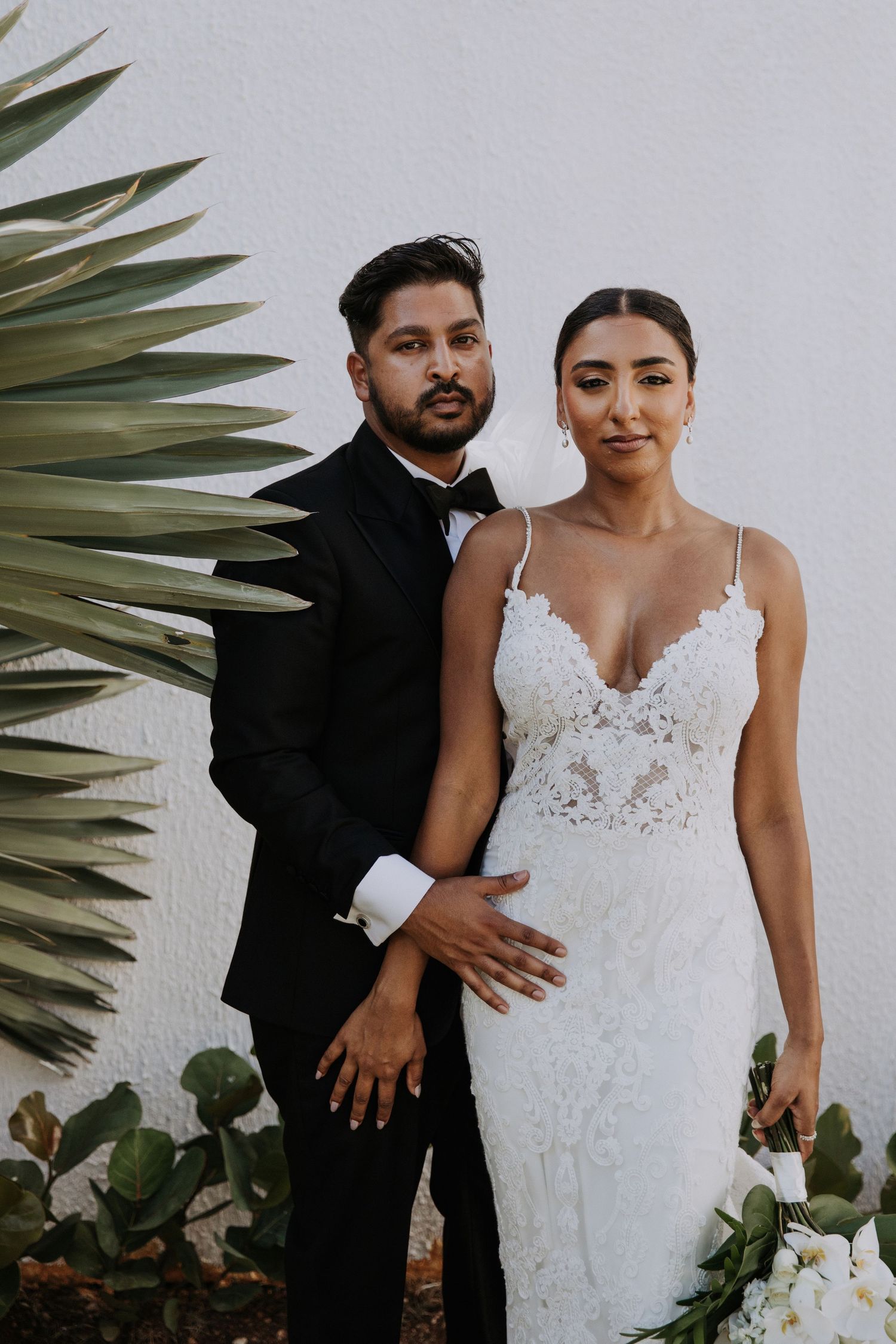 Intimate wedding portraits captured against white wall with lush palm leaves as natural decor.