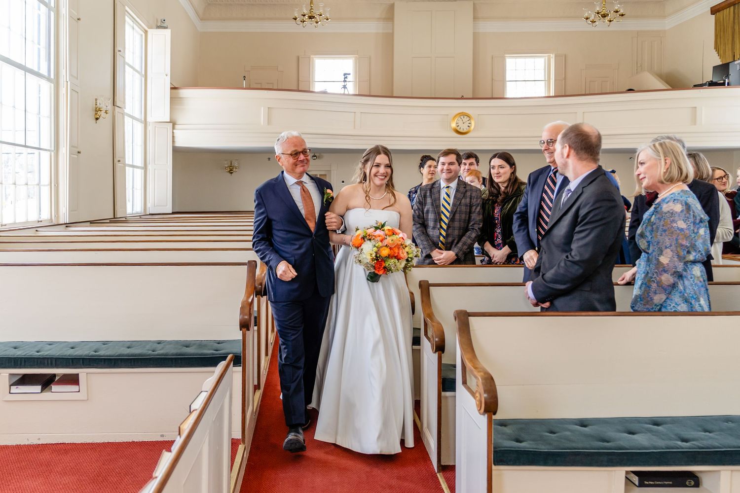 Wedding party gathers on wooden church pews during bright indoor ceremony while natural light streams through windows.
