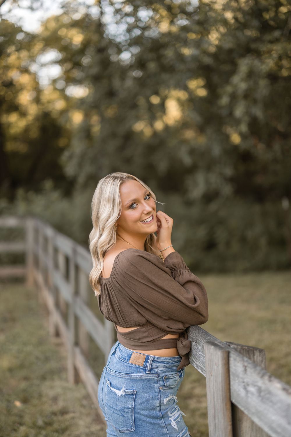 Person in brown sweater and jeans poses casually against rustic wooden fence during golden hour outdoors.