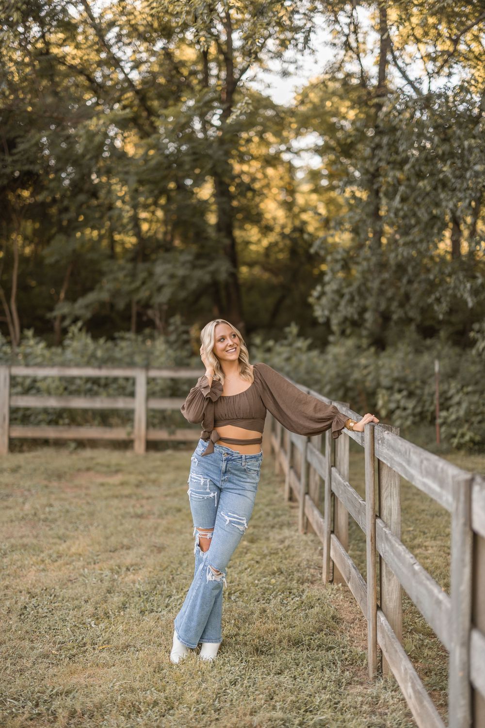 A woman in casual attire poses along a wooden fence at sunset in a rustic countryside setting.