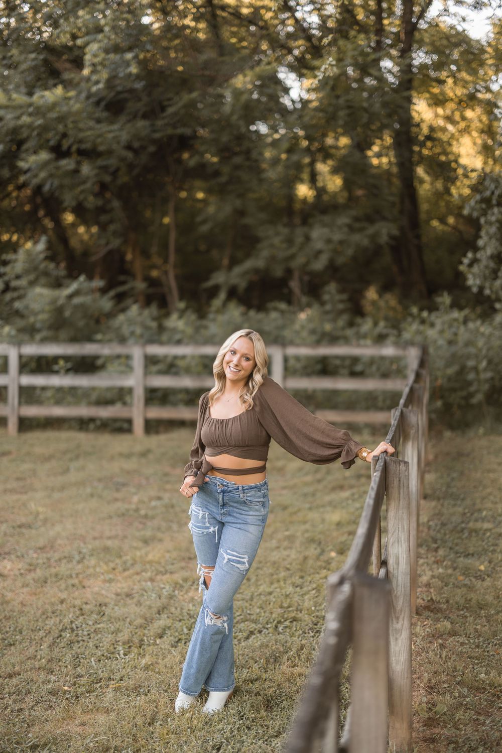 Person in a brown crop top and distressed jeans poses casually by a rustic wooden fence at golden hour in a pastoral setting.