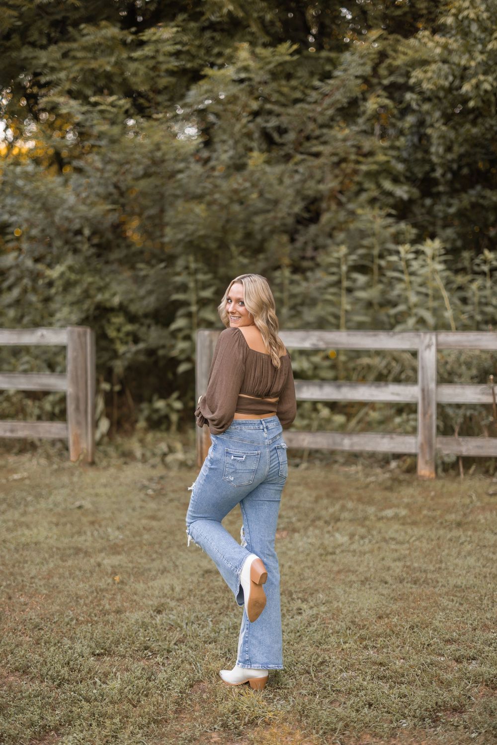 Person in casual wear poses playfully near a wooden fence on a grassy field during a golden hour photoshoot.