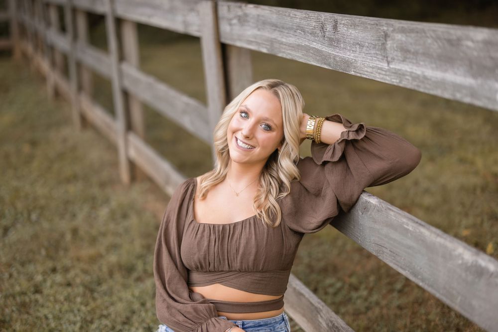 A young woman in a brown crop top and jeans poses casually by a wooden fence on a grassy field.
