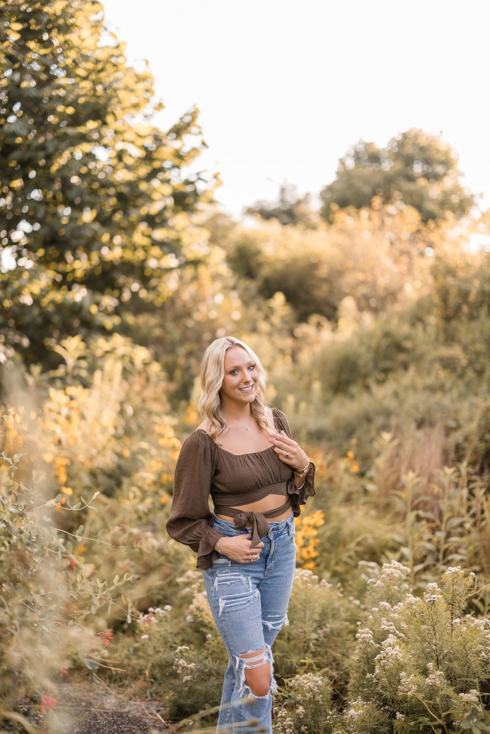 Golden light illuminates a person wearing jeans and brown top posing in a field of tall wildflowers at sunset.
