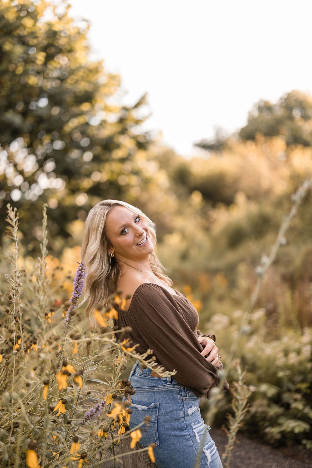 A woman laughs joyfully in a brown sweater and jeans amidst golden wildflowers during sunset.