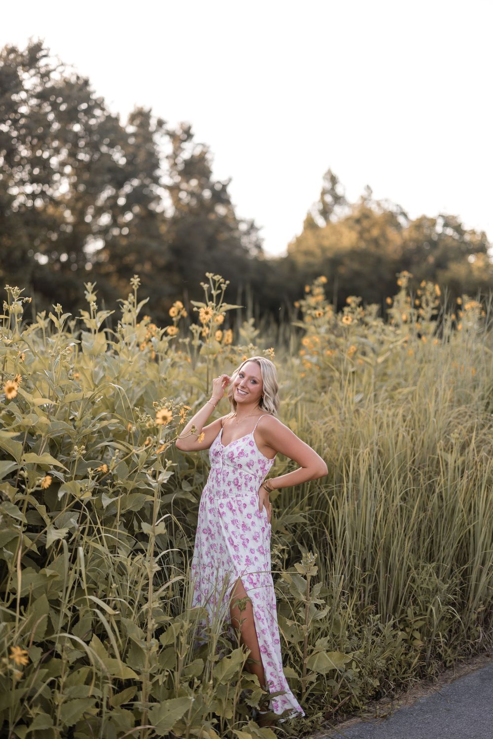 A woman in a floral dress poses beside tall sunflowers during golden hour on a rural path.
