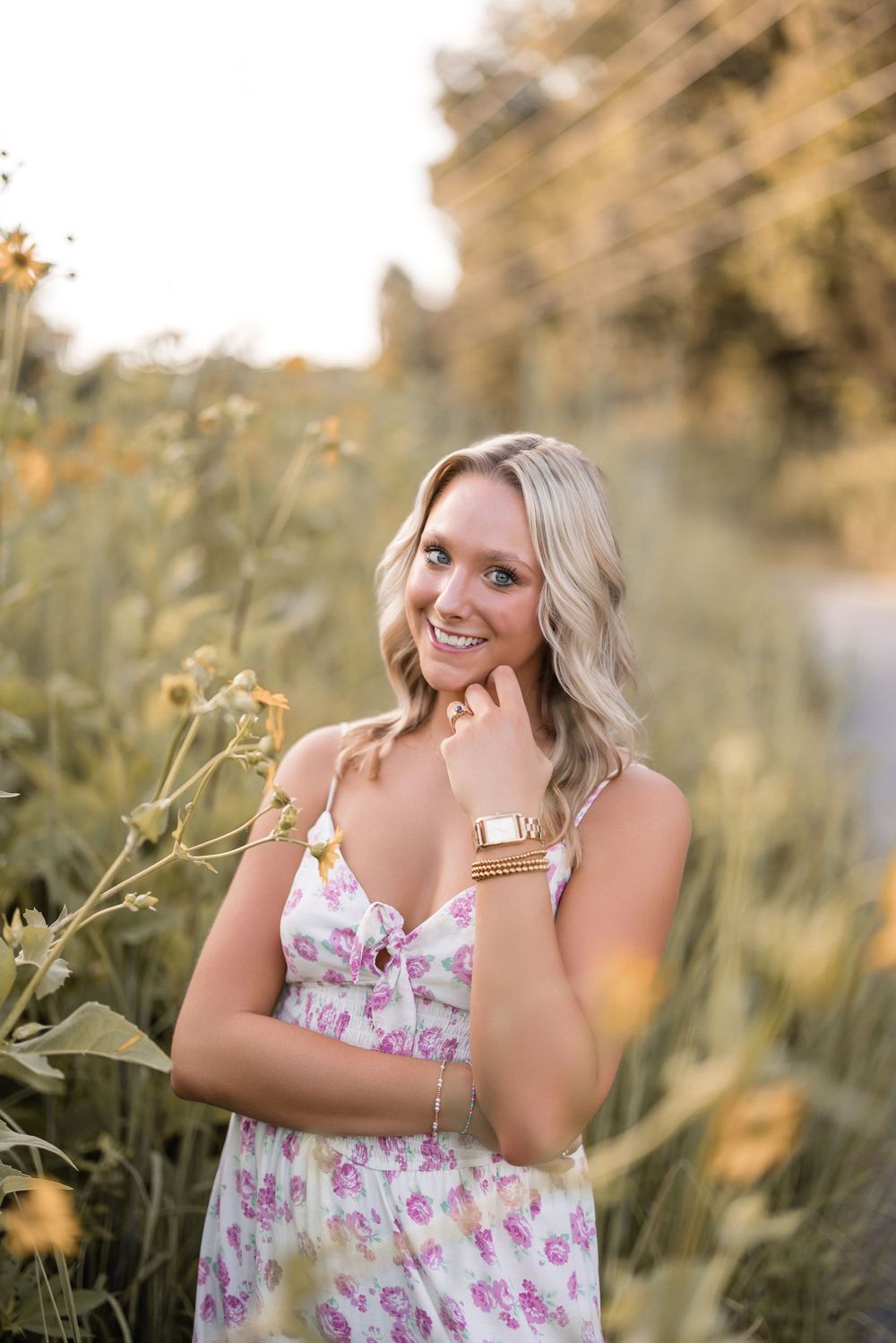 A person in a floral sundress poses outdoors among wildflowers at golden hour in a natural setting.