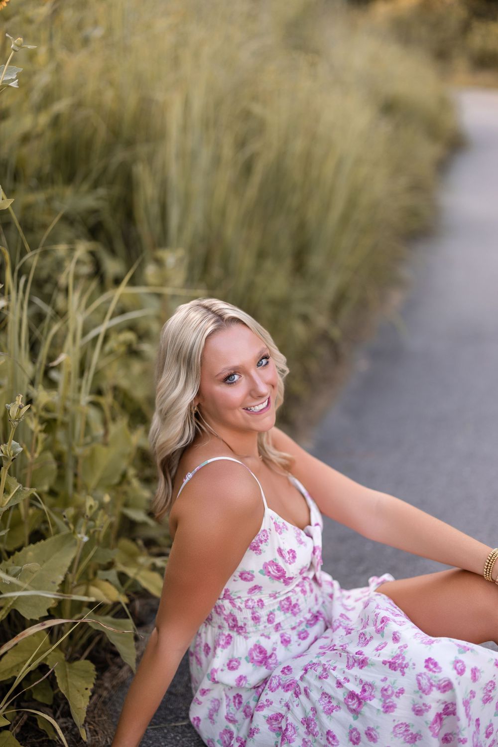 A woman in a floral summer dress poses playfully on grass near a path during golden hour.