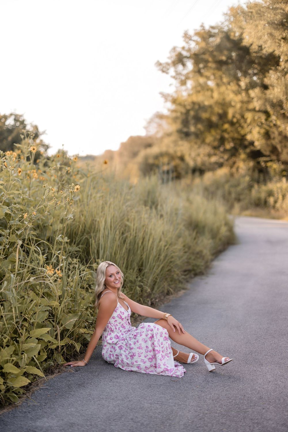 A person in a floral dress sits on a curved pathway surrounded by tall grasses and greenery at sunset.