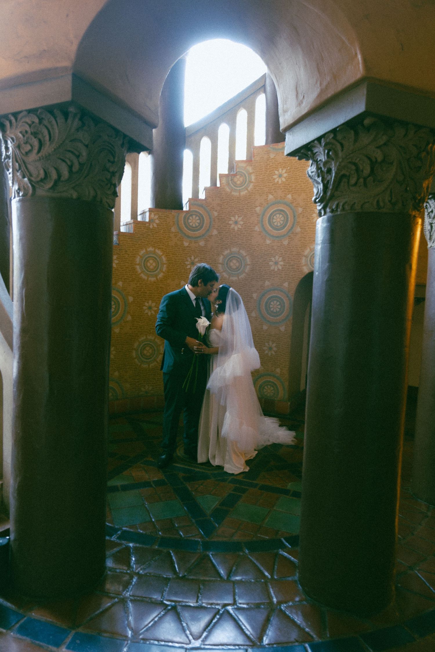 A couple in wedding attire share an intimate moment framed by ornate columns and arched windows in a Spanish-style venue.