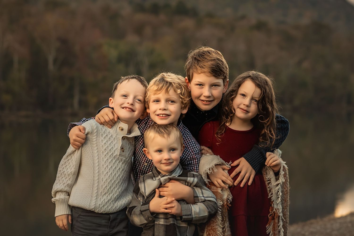 A group of children wearing cozy sweaters smile together by a lake during golden hour.