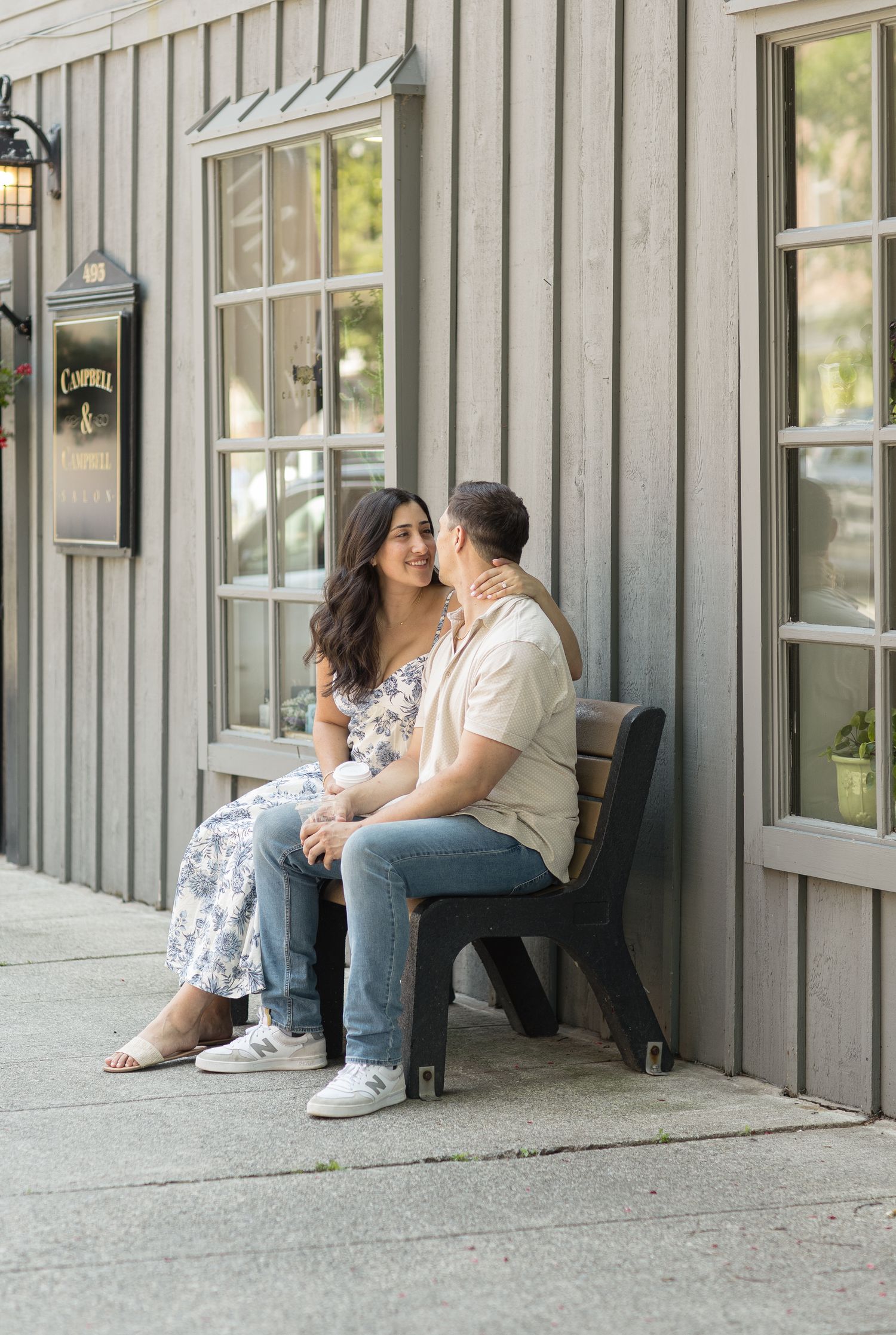 A couple sits lovingly together on a rustic wooden porch bench outside a gray building with windows.