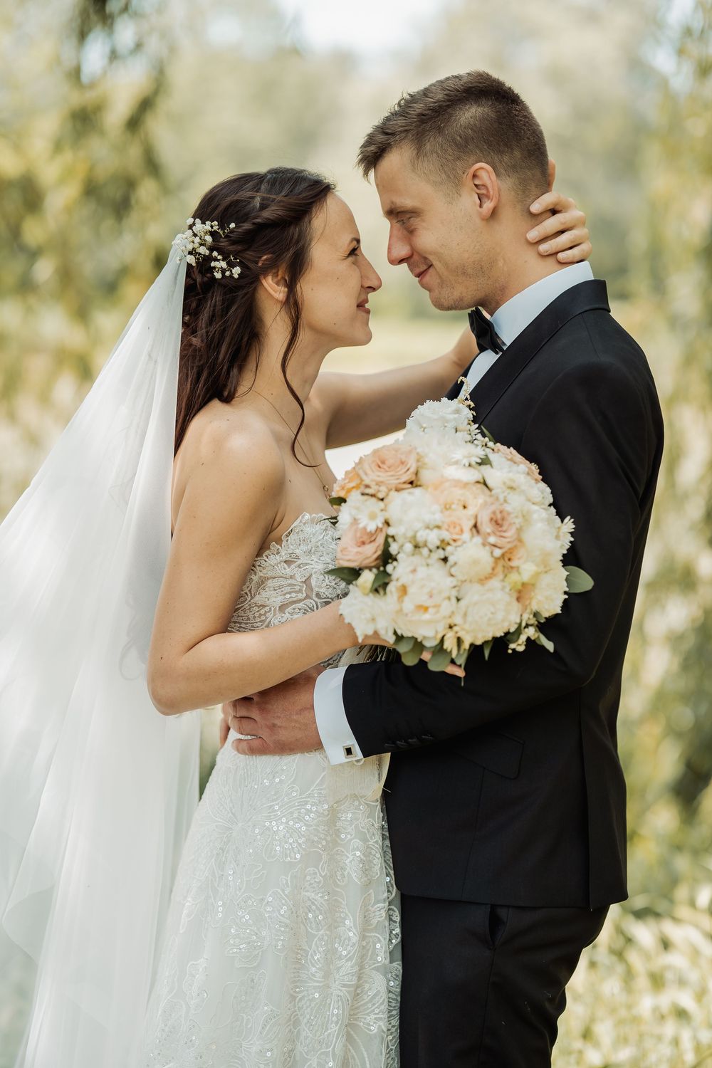 Romantic outdoor wedding portrait of newlyweds embracing with peach and white floral bouquet against blurred nature background.