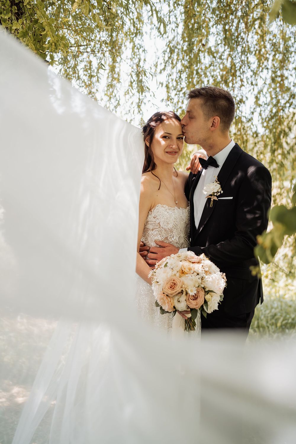 A romantic wedding photo under a weeping willow tree with a peach rose bouquet and elegant attire.