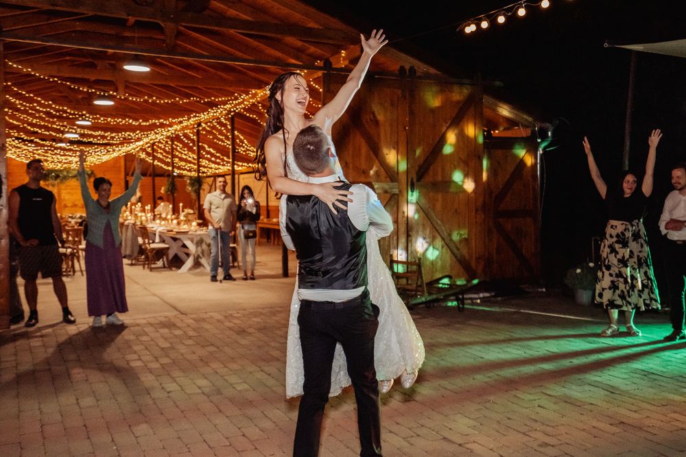 A couple dances together at a rustic wedding reception venue decorated with warm string lights and greenery.