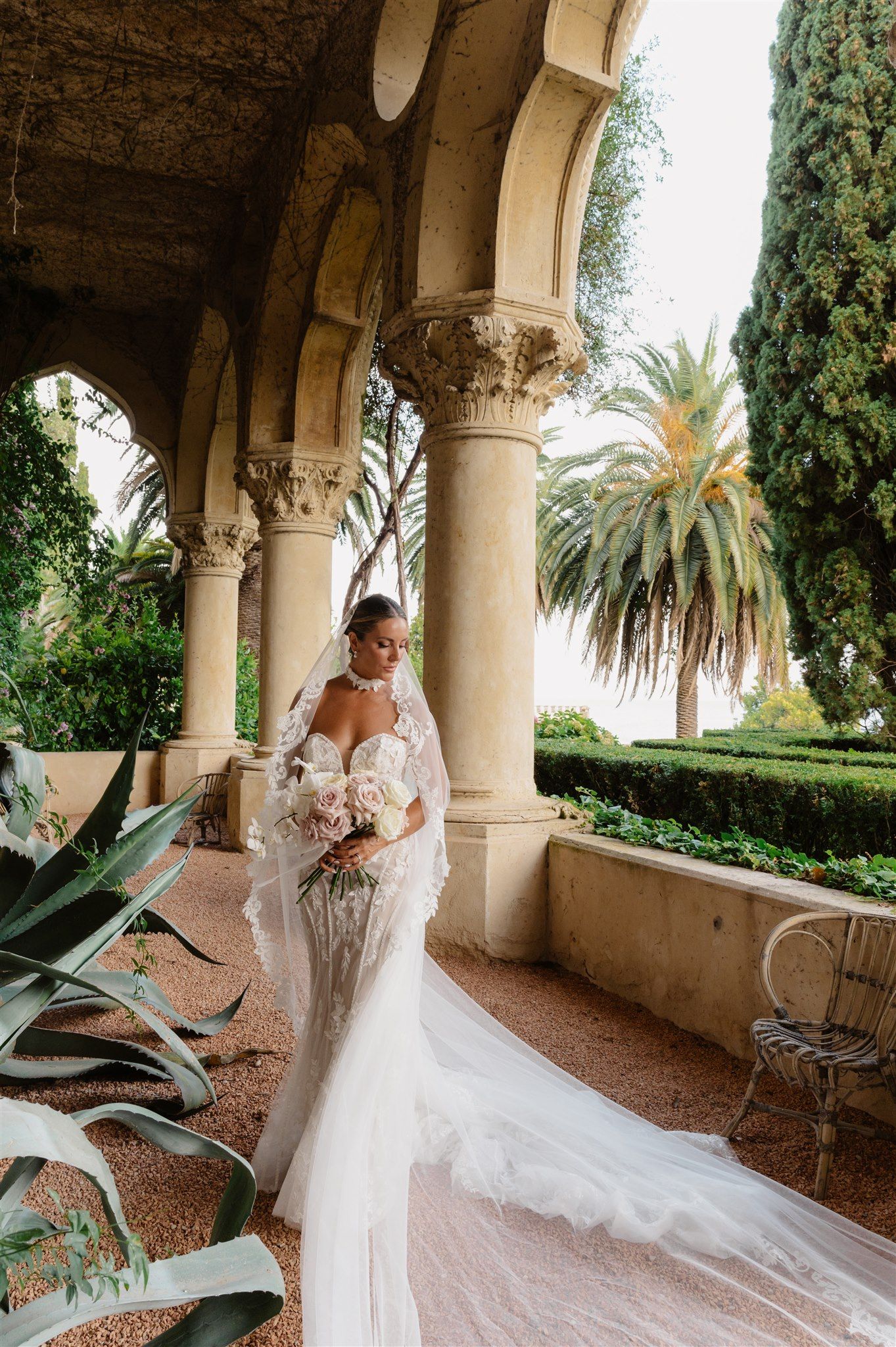 Bride in lace gown with flowing train poses on Mediterranean villa terrace with palm trees.