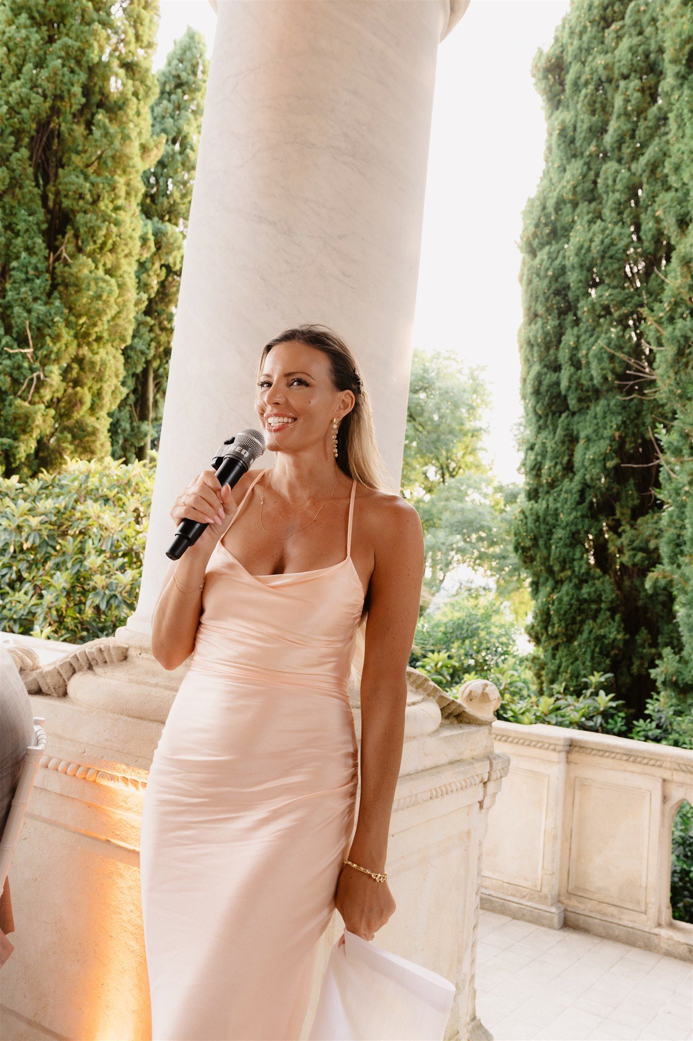 Woman in blush slip dress speaks into microphone on elegant stone terrace with cypress trees.