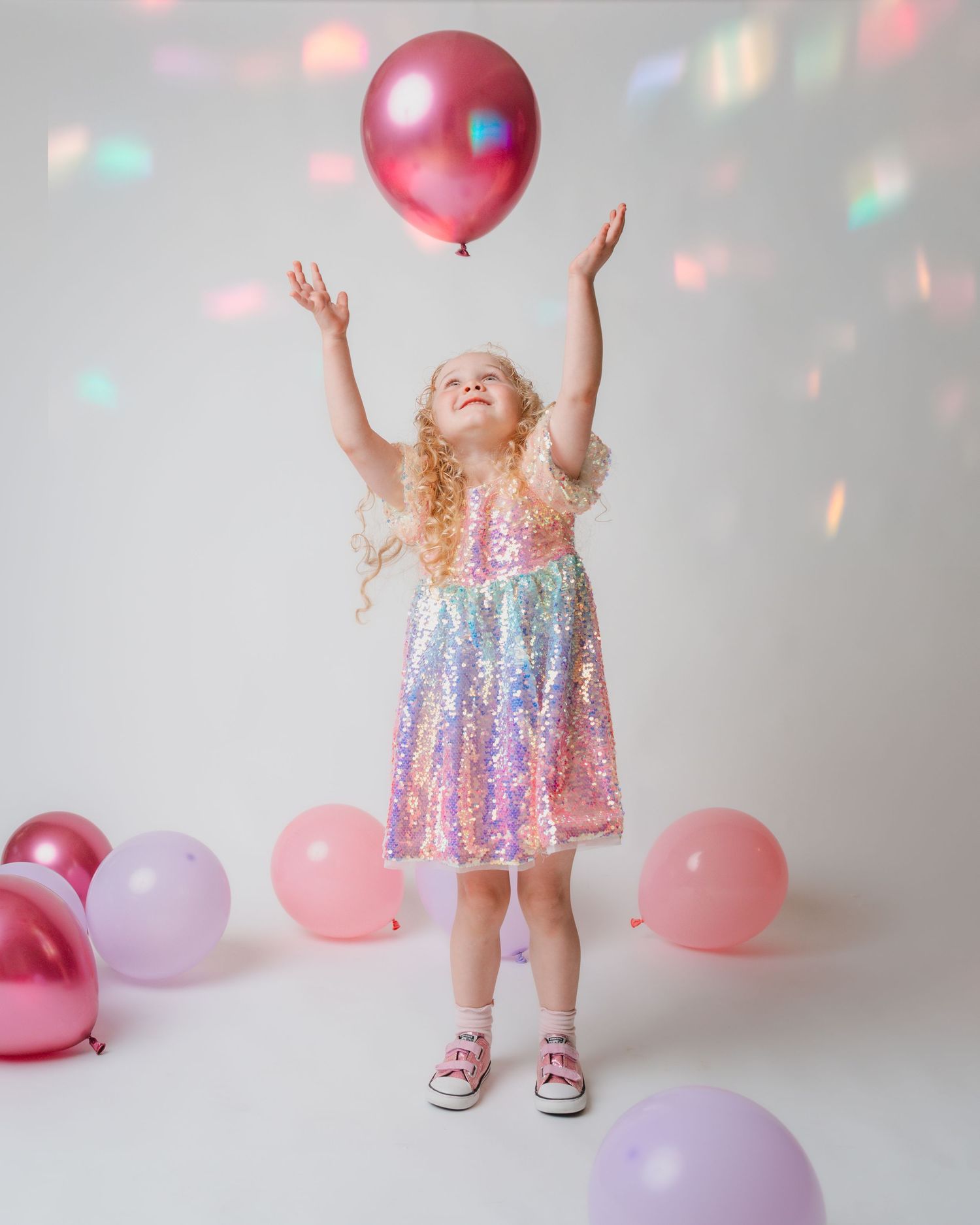 Young girl in a rainbow sequin dress joyfully tossing a pink balloon surrounded by colorful balloons.