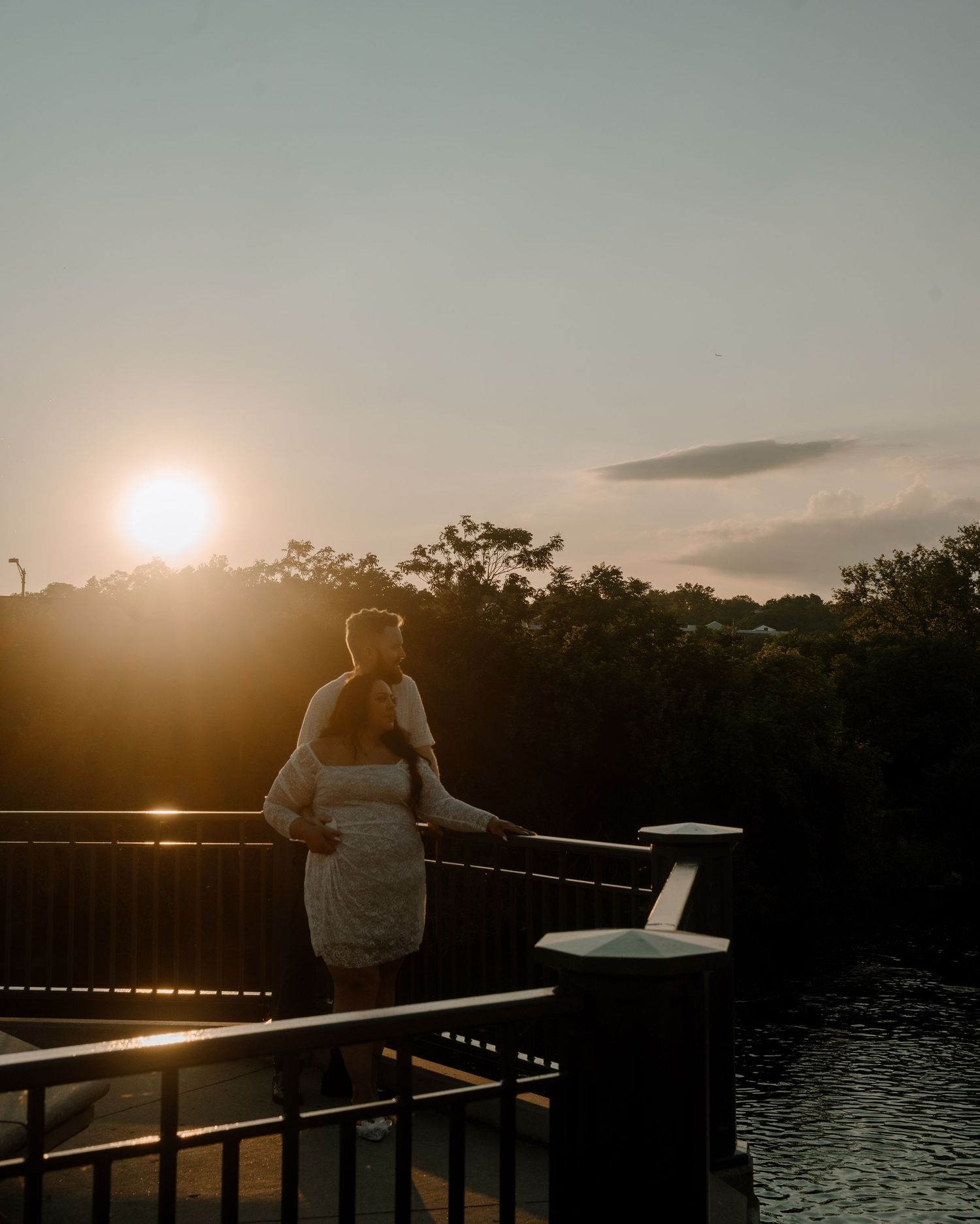 A silhouette watches the golden sunset over a waterfront railing with trees and reflective water in the foreground.