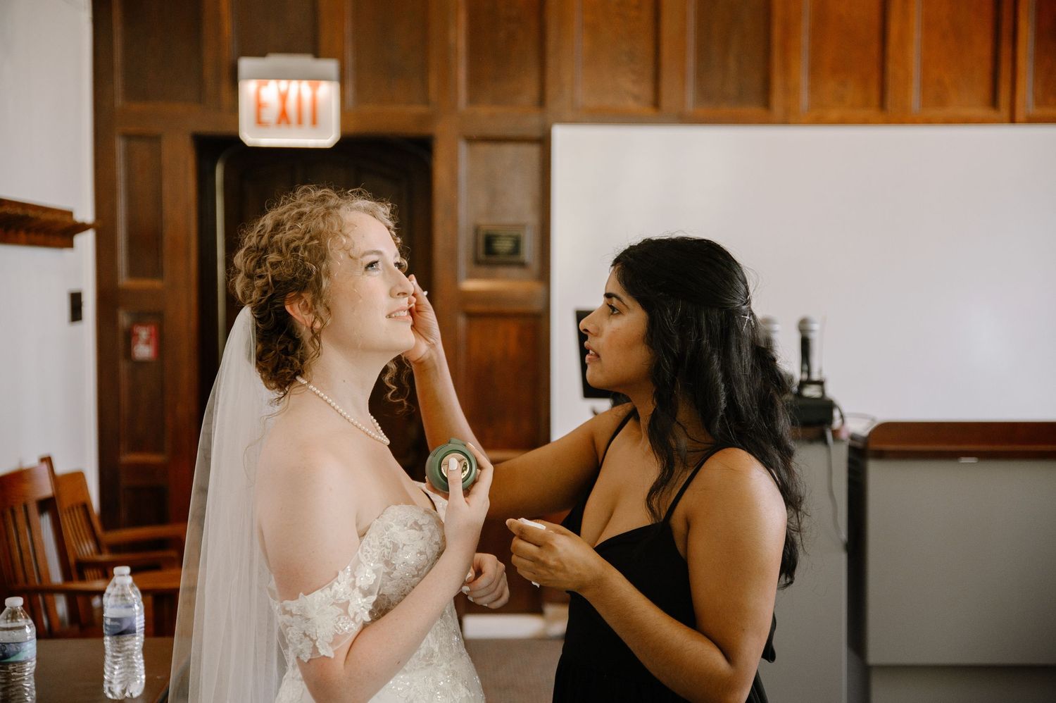 Final touchups being applied to a bride wearing a lace wedding gown and veil before the ceremony.