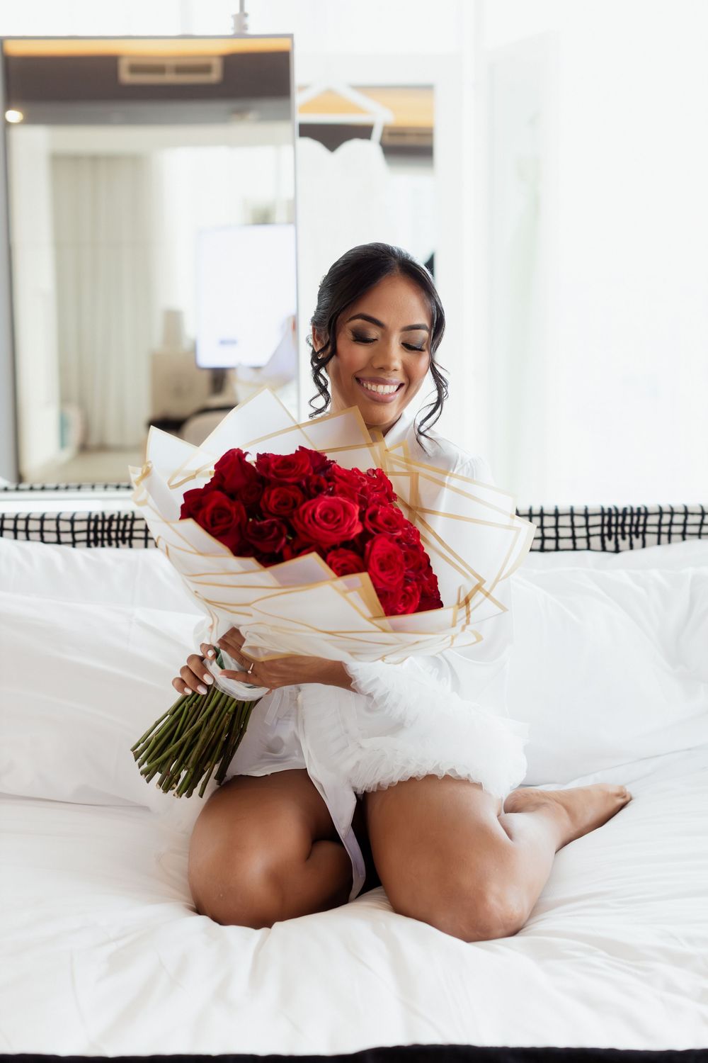 A bride in white dress relaxes on a bed holding a vibrant red rose bouquet.
