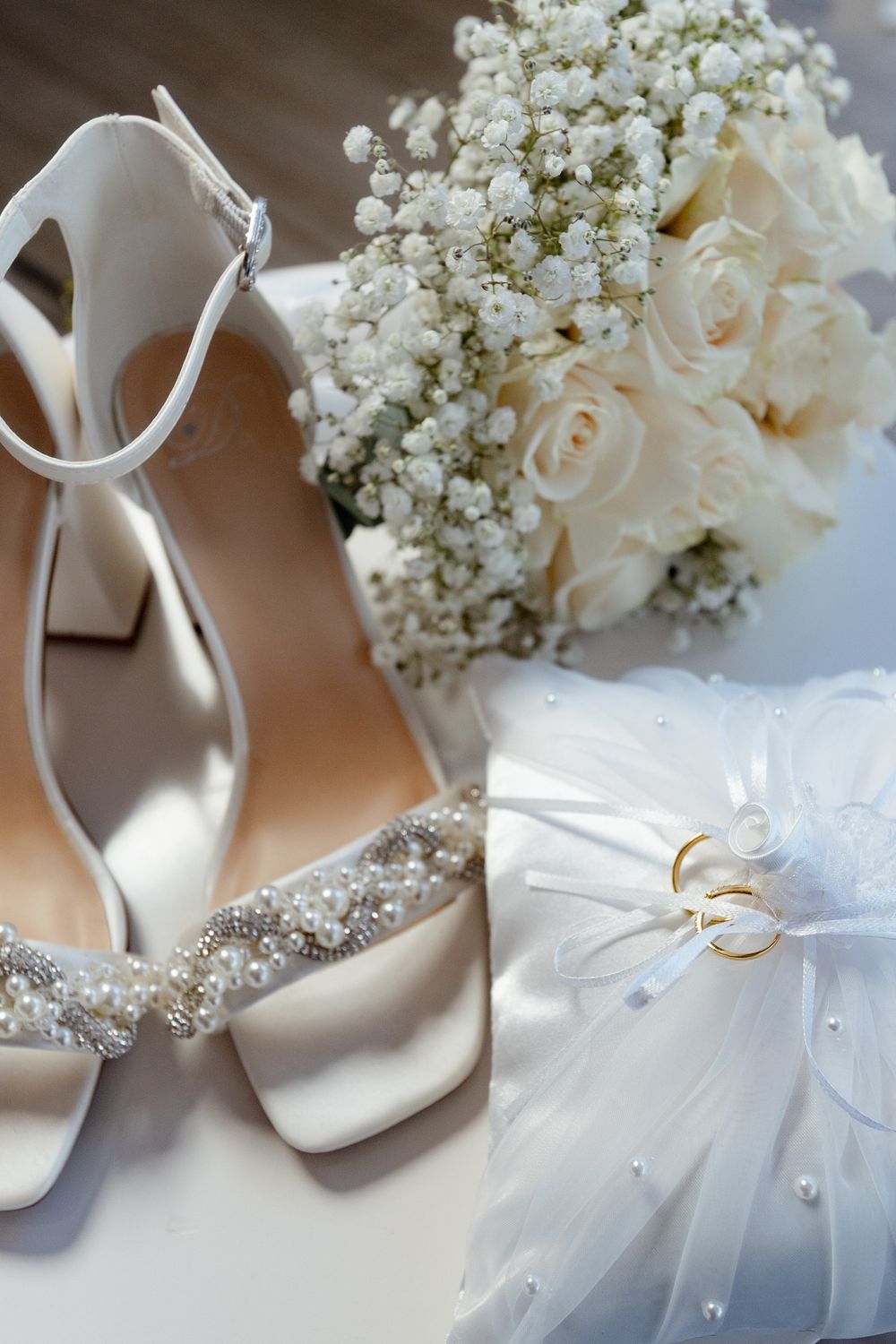 Elegant white bridal shoes with crystal embellishments next to a delicate bouquet of white roses and baby's breath.