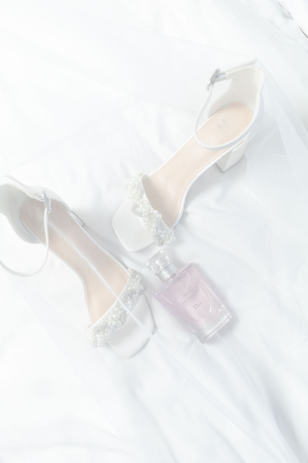 Elegant white bridal shoes with lace details paired with a delicate perfume bottle on white fabric background.