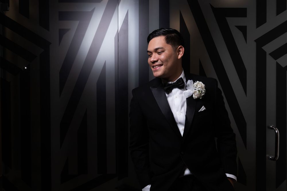 A groom in a classic black tuxedo and white boutonniere smiles while standing against a geometric patterned wall.