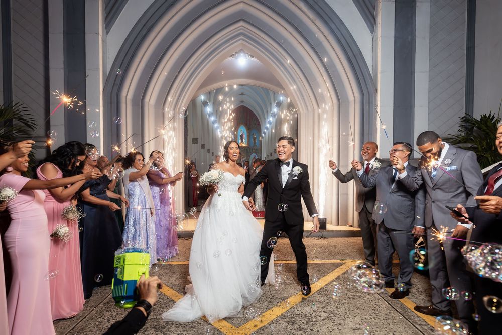 Newlyweds celebrate and dance down the aisle of a grand cathedral with arched ceilings and wedding guests cheering.