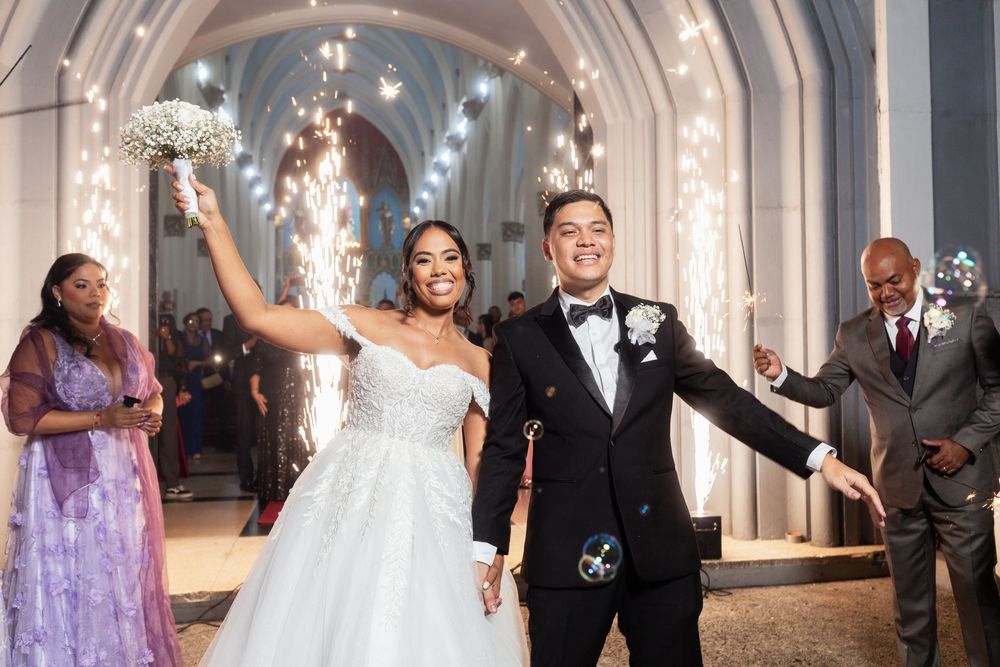 Wedding party wearing black tuxedos and lavender dresses celebrates in an elegant white ballroom with marble columns.
