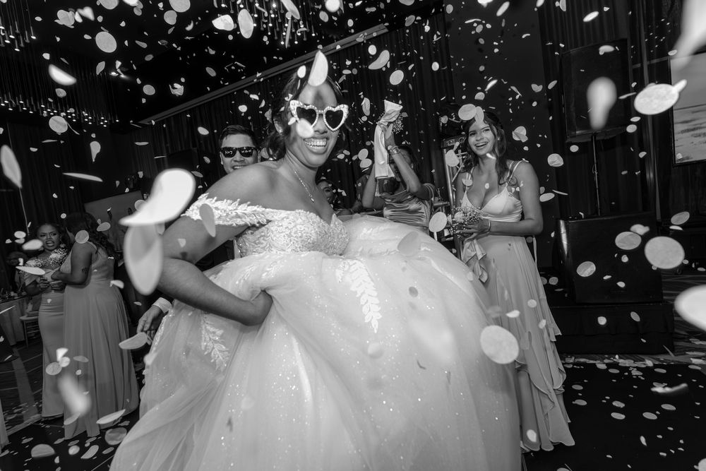 Black and white photo of a wedding celebration with white petals falling and guests in formal attire dancing in the background.