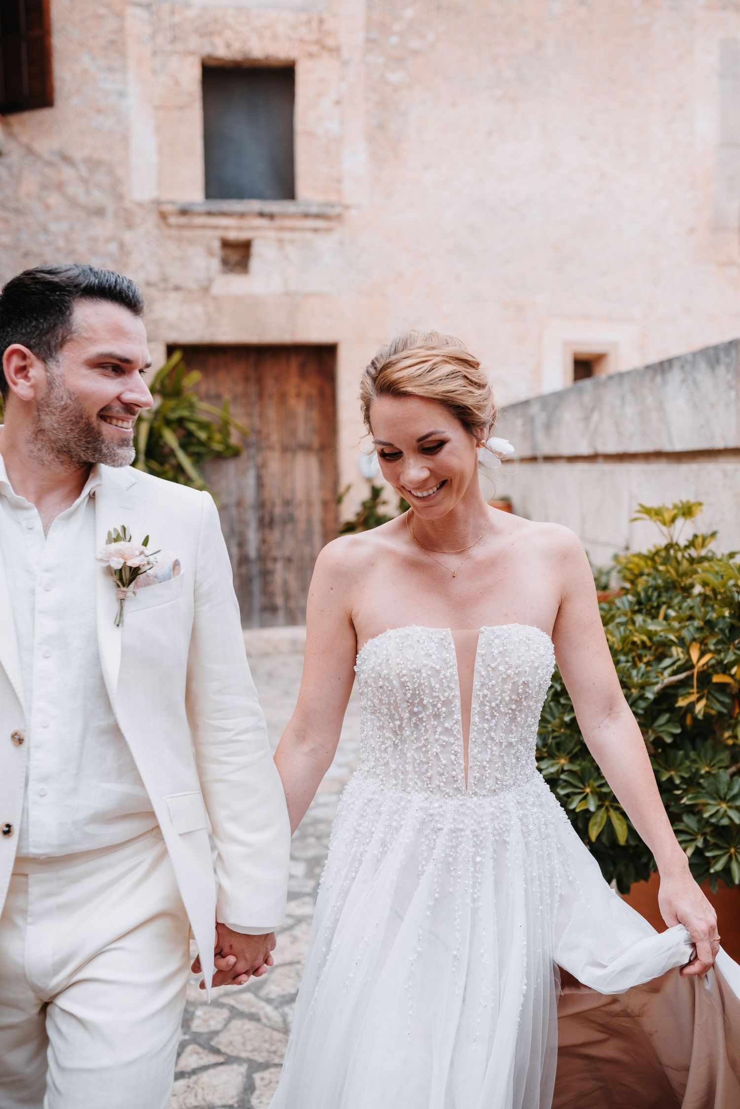 Ein fröhliches Paar in weißer Hochzeitskleidung lacht gemeinsam im mallorquinischen Innenhof der Finca Xiclati. Er schaut sie verliebt an.
