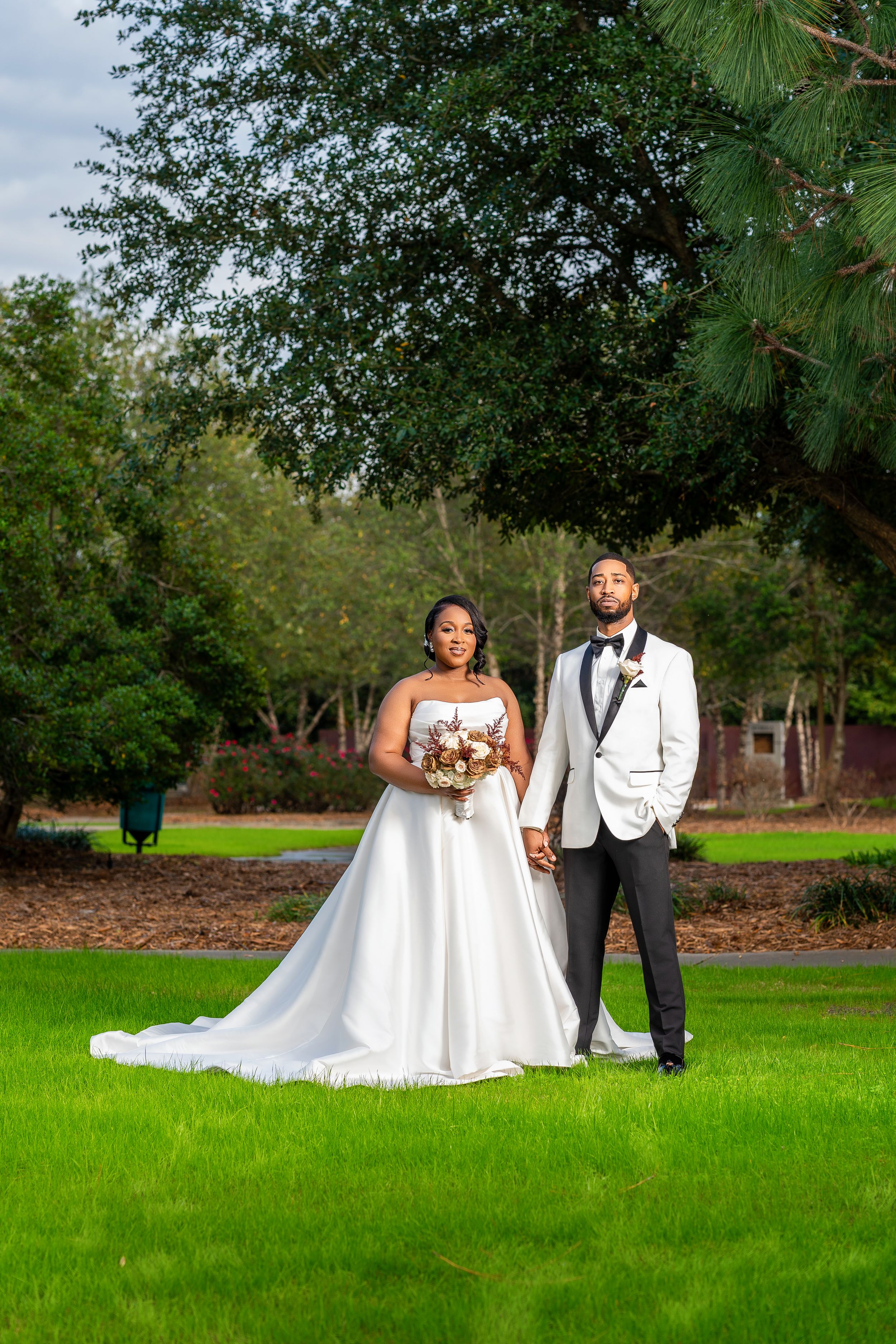 A wedding couple stands together on a lush green lawn with trees and foliage in the background.