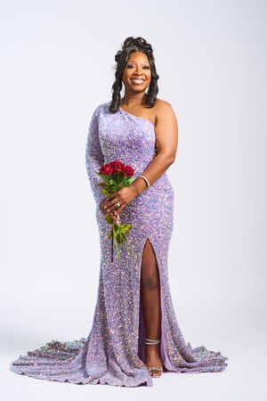A model in a sparkly lavender gown poses against a white backdrop holding a vibrant red bouquet of flowers.