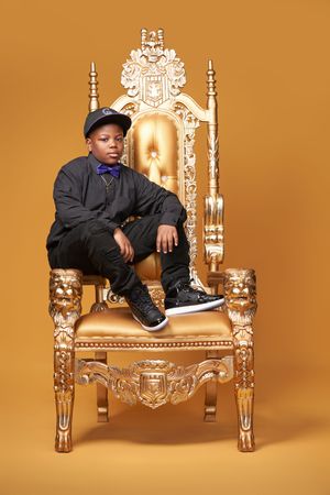 Student seated casually on ornate gold throne chair with carved designs against orange background.