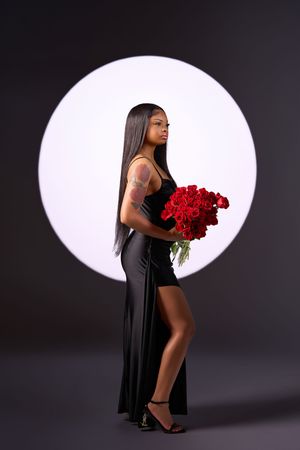 Elegant model in black dress poses with vibrant red roses against dark background with circular white spotlight.
