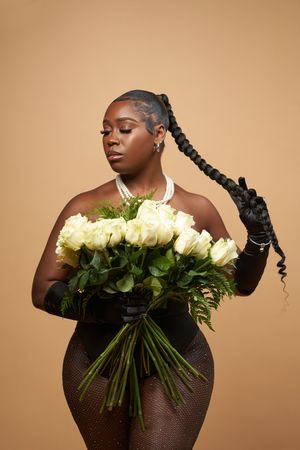 Elegant fashion portrait with long braided hair, black gloves and dress, holding a bouquet of white roses against beige background.