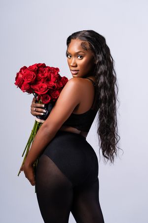 Figure in black athletic wear poses with red roses against light background while looking over shoulder.